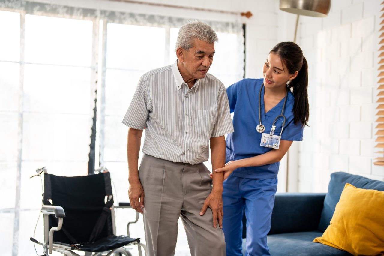 An elderly man receiving assistance from a nurse to stand up in a bright, comfortable room with a wheelchair nearby.