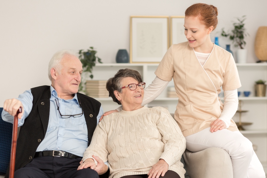 An elderly couple sitting together on a couch, smiling, while a caregiver gently interacts with them.