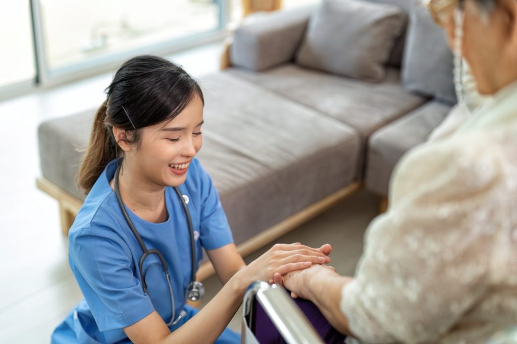 A nurse smiling and gently holding the hand of an elderly woman in a bright, cozy room.