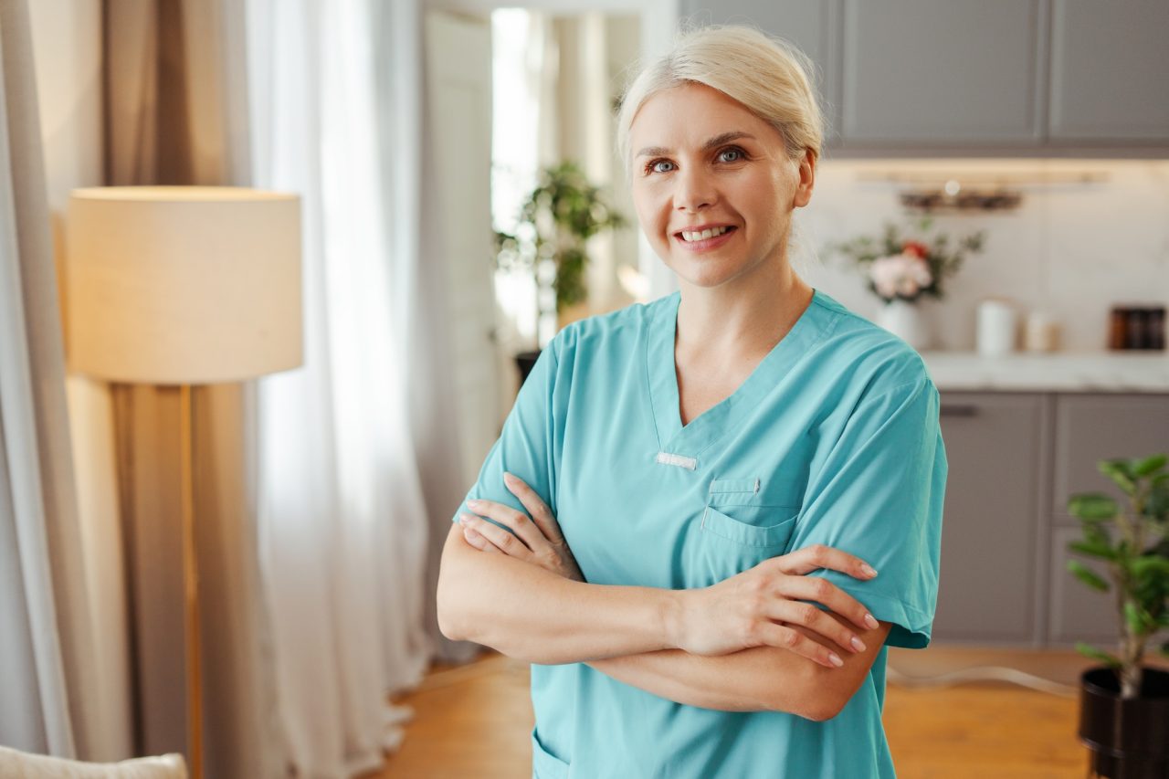 A smiling female nurse in teal scrubs standing with her arms crossed in a well-lit room.