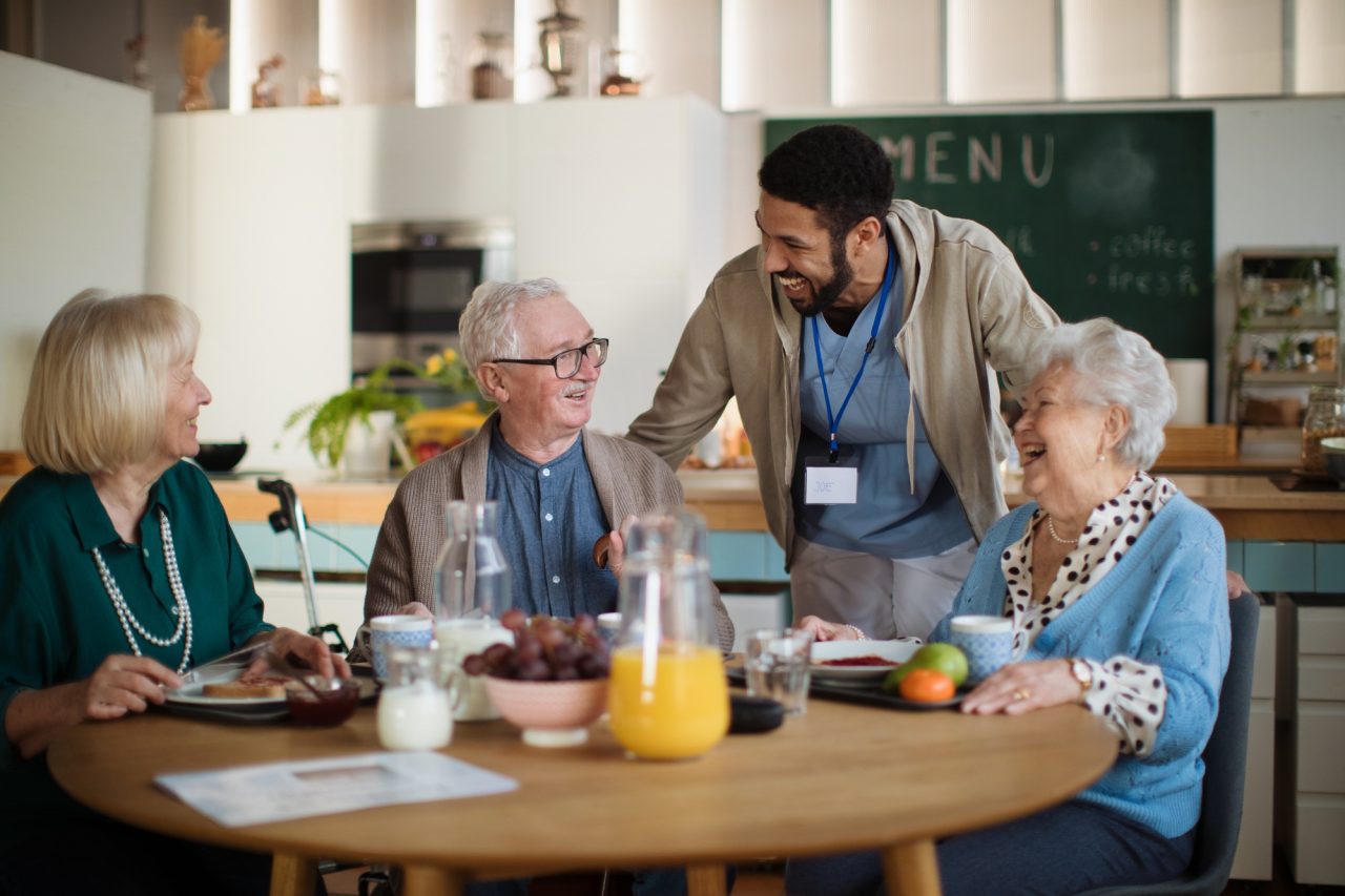 A caregiver chatting and laughing with elderly individuals over breakfast at a kitchen table.