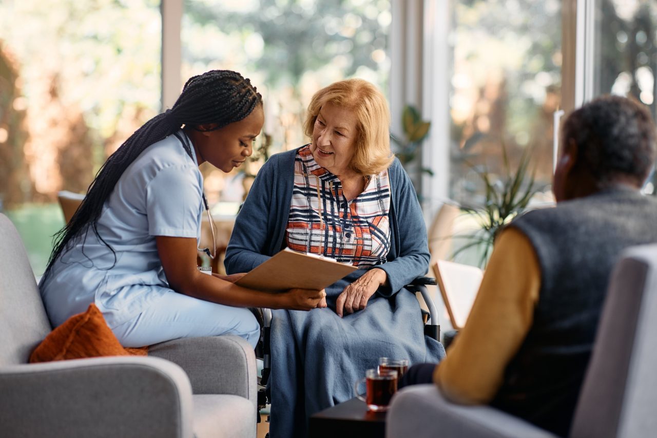 A nurse showing a document to an elderly woman while another person sits nearby, all smiling.