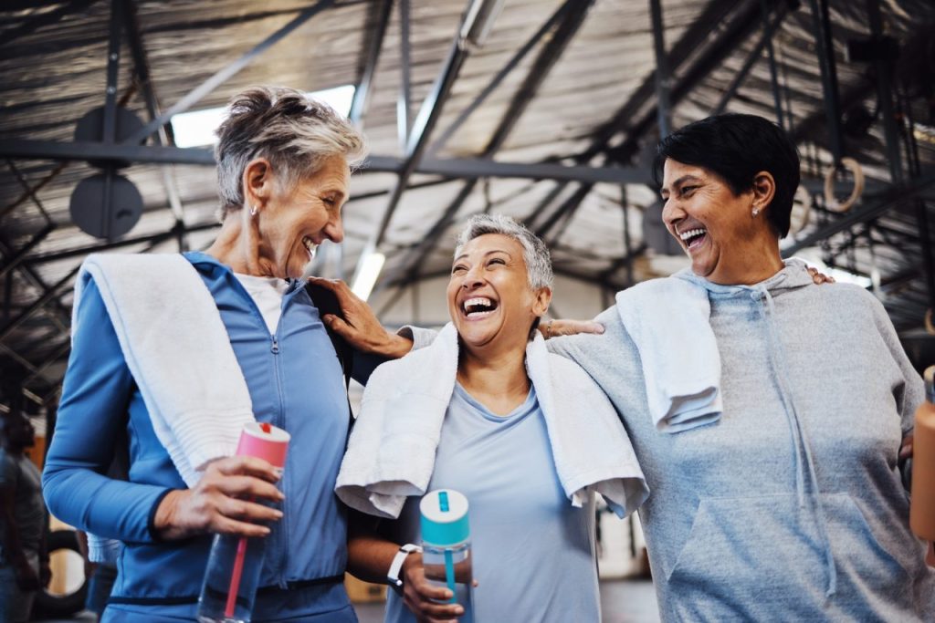 Three women laughing together after a workout, each holding a water bottle.