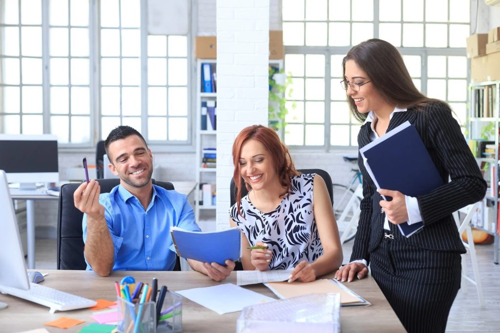 Three colleagues smiling and discussing documents in a bright office setting.